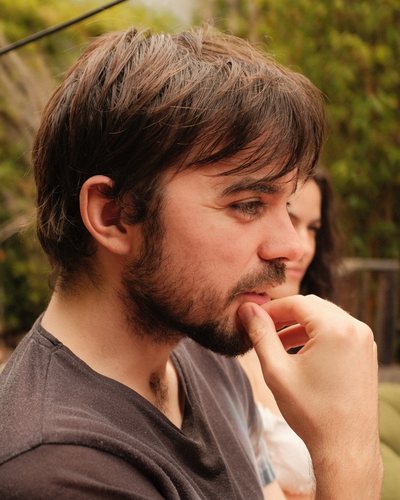 Cormac outdoors in profile, fingers resting at his mouth, looking at something off to the side