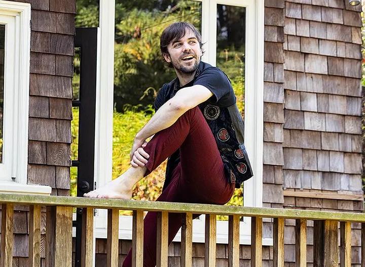 Cormac sitting on the railing of a wooden back deck, one bare foot up, laughing at something off to the side